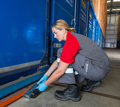 Junge Frau in Arbeitskleidung und Handschuhen platziert einen kleinen, schwarzen Kasten am Boden eines blauen Containers.