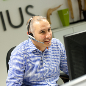 A smiling white man wearing a checked shirt and a headset is talking on the phone and looking at a screen.