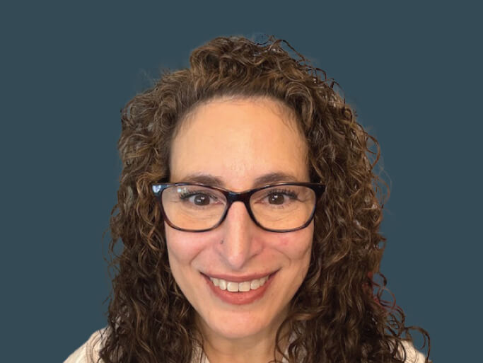 A portrait of a smiling middle-aged woman with brown curls and glasses against a blue-grey background.