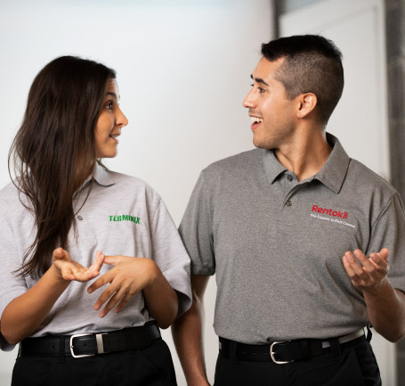 A female employee in a light grey 'Terminix' polo shirt and a male employee in a dark grey 'Rentokil' polo shirt are standing next to each other, engaged in a friendly conversation. They are both gesturing with their hands as they speak.
