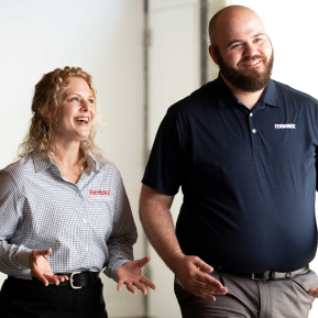 Two smiling employees, a man in a navy blue Terminix shirt and a woman in a checked Rentokil shirt, chat and walk in a hallway.