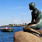 Copenhagen, Denmark - The Little Mermaid bronze statue by Edvard Eriksen, with sightseeing boats in the background.