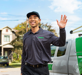 A smiling male Terminix employee in a grey shirt and black cap is waving while standing in front of a Terminix truck.