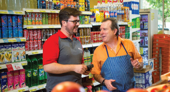 A Rentokil employee in a gray shirt and a store clerk wearing an apron are talking in an aisle between two shelves.
