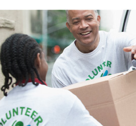 A smiling man wearing a T-shirt that says “Volunteer” hands a large cardboard box to another person who is also wearing a T-shirt that says “Volunteer.”