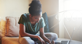 A young woman sits on a couch, studying and taking notes. An open laptop lies in front of her.
