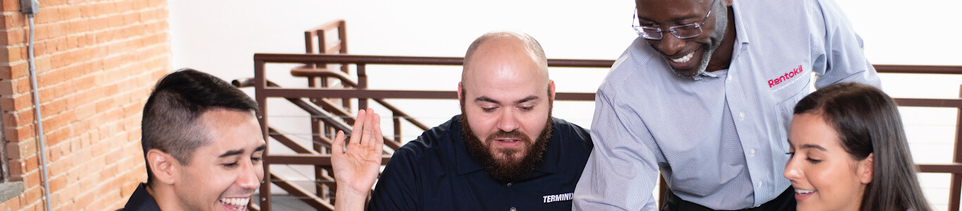 A friendly male Rentokil service technician wearing a company uniform smiles at the camera. He is dressed in a dark blue polo shirt with a red collar and the Rentokil logo on the chest, and dark trousers. He is standing outdoors in a professional pose.