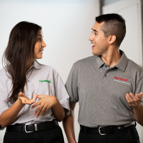 A smiling male Rentokil employee and a female Terminix employee are standing in grey shirts and talking.
