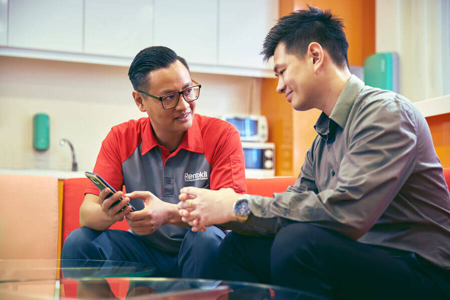 A **Rentokil** technician in a red uniform is sitting on a sofa and showing something on his phone to a smiling man.