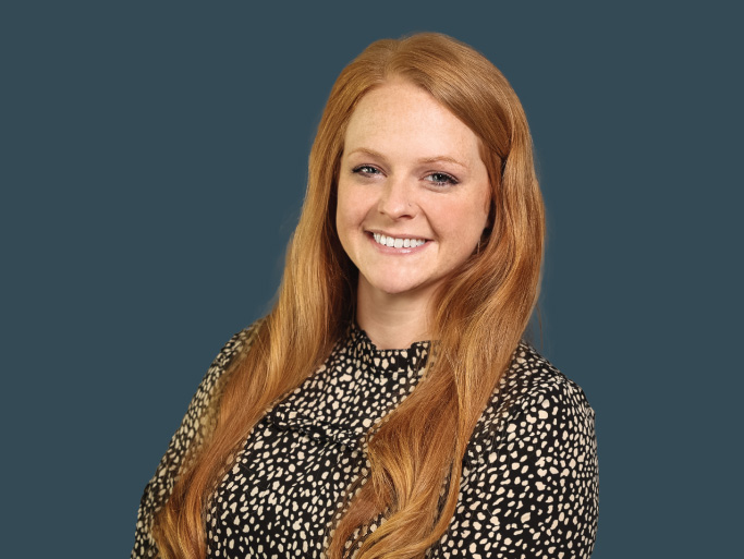 A portrait of a smiling young woman with long red hair wearing a leopard-print blouse against a blue-grey background.