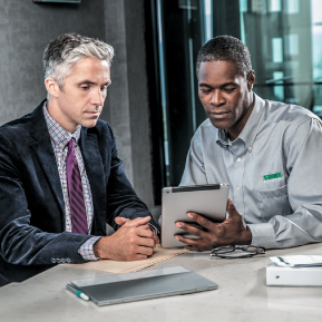 Two men are sitting at a table, and one man in a gray Terminix shirt is showing something on a tablet to another man in a suit.