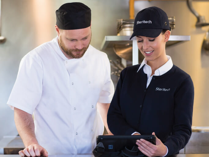 A Steritech consultant in a dark blue sweater and a hat shows a tablet to a chef in a kitchen.