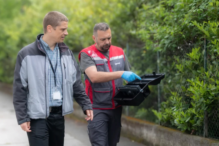 Dos hombre caminan juntos, mientras uno de ellos, técnico de Rentokil, enseñando una trampa de cebo para ratas