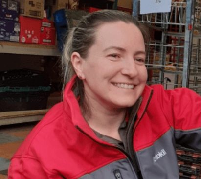 A woman with light hair tied back, wearing a red and gray work jacket, smiling and is near shelves stocked with boxes and items.