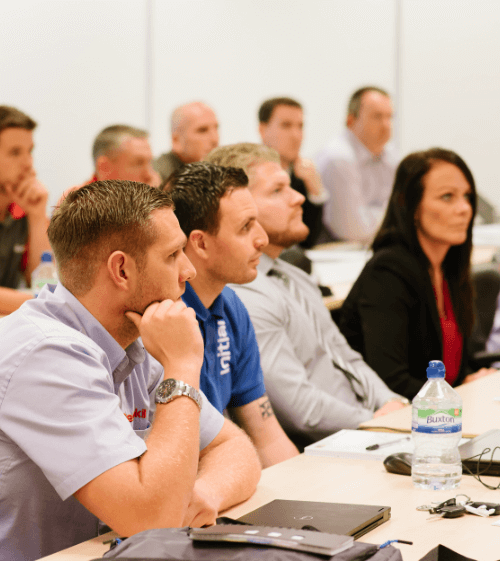 A group of people, both men and women, sit at tables in a meeting room.