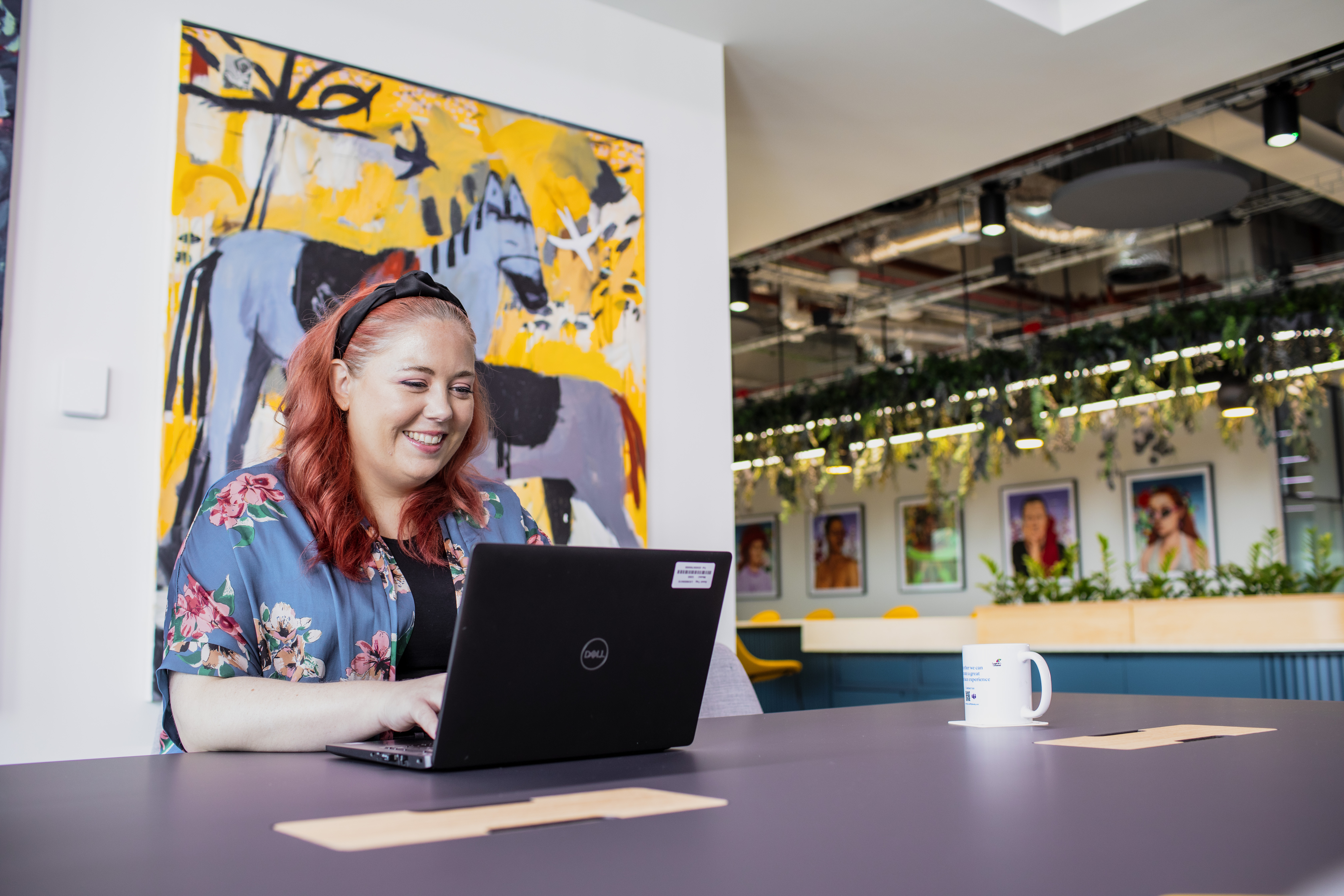 A woman with red hair and a floral top on a laptop in a colourful office