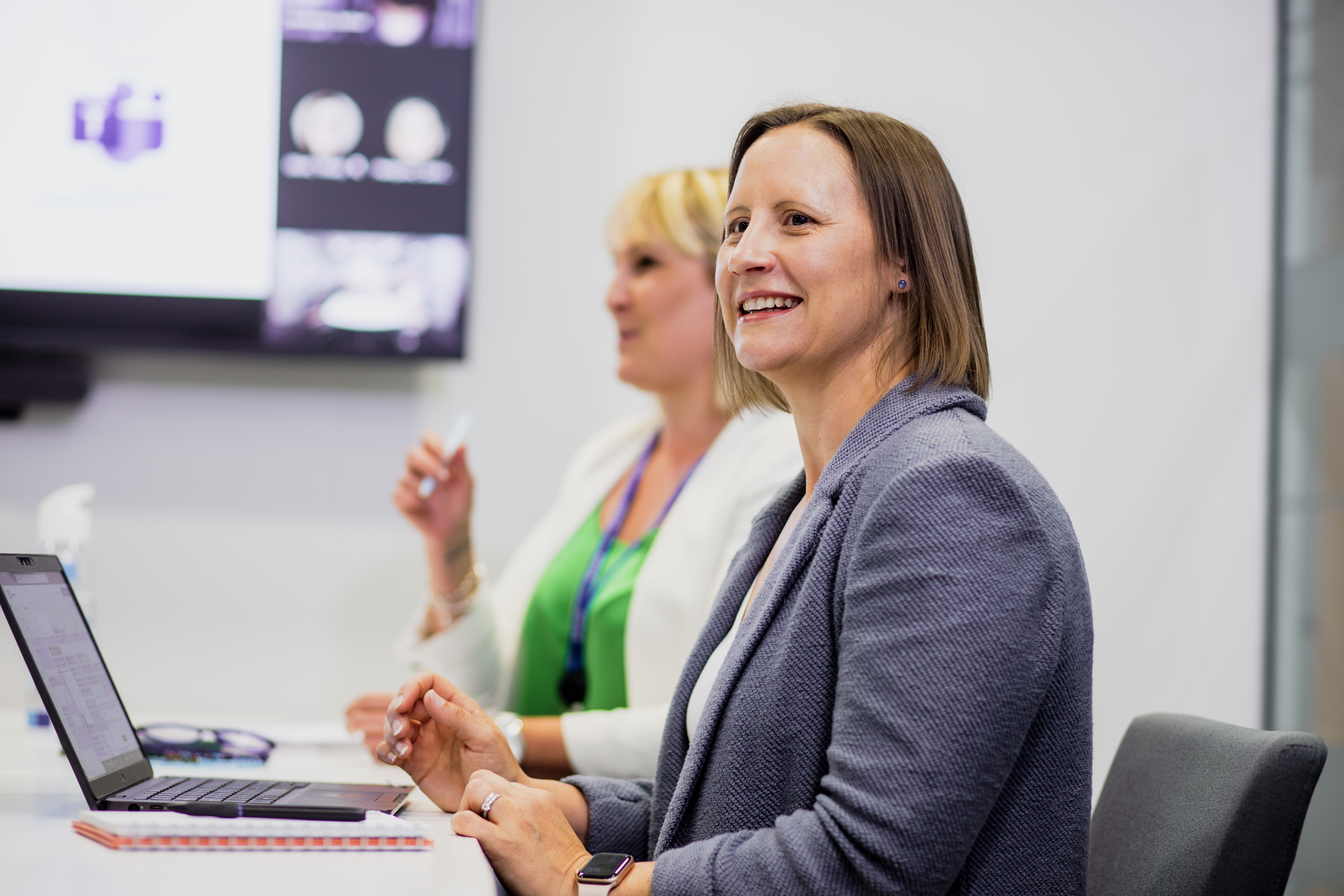 Two women at a desk on a laptop in a work meeting in the office with a big screen behind them.