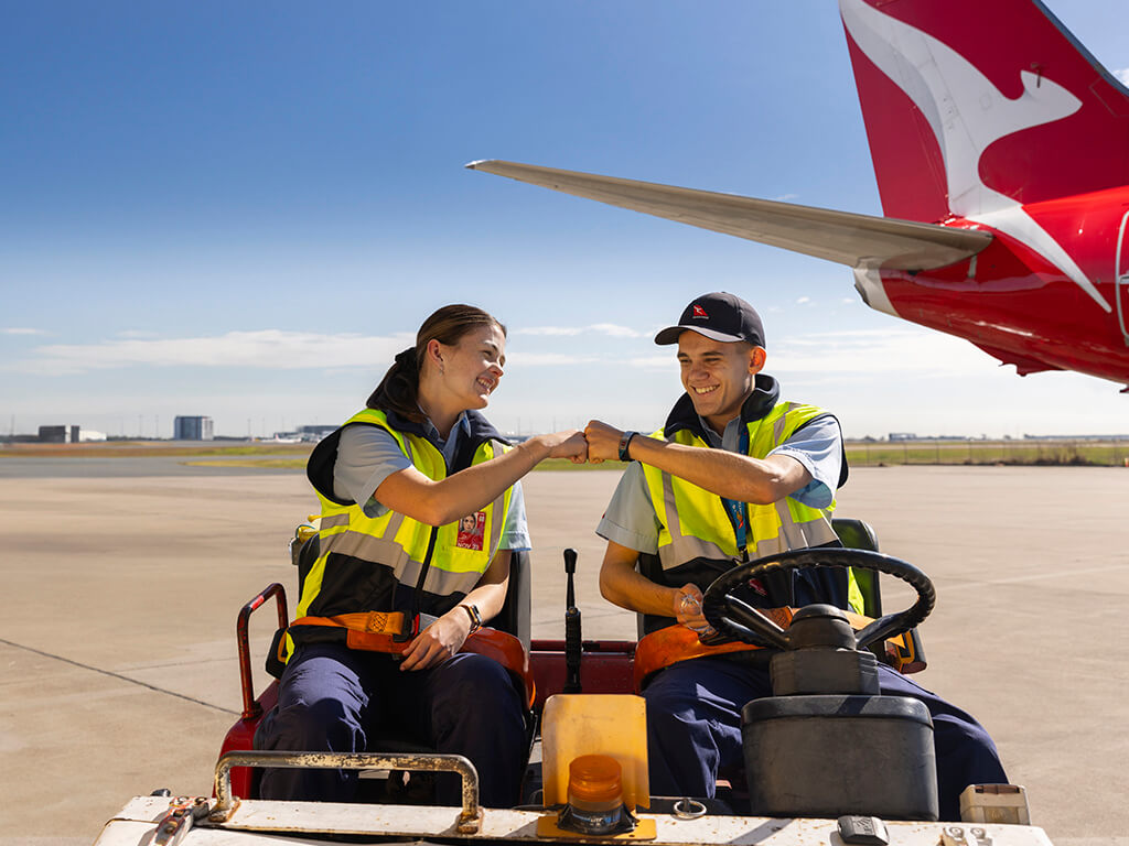 Two Qantas Apprentice Engineers smiling and operating a tug on the tarmac next to an aircraft