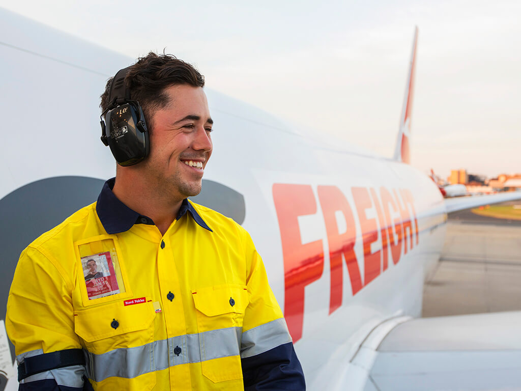 Smiling Qantas Freight employee on the tarmac beside an aircraft, wearing earmuffs and safety gear