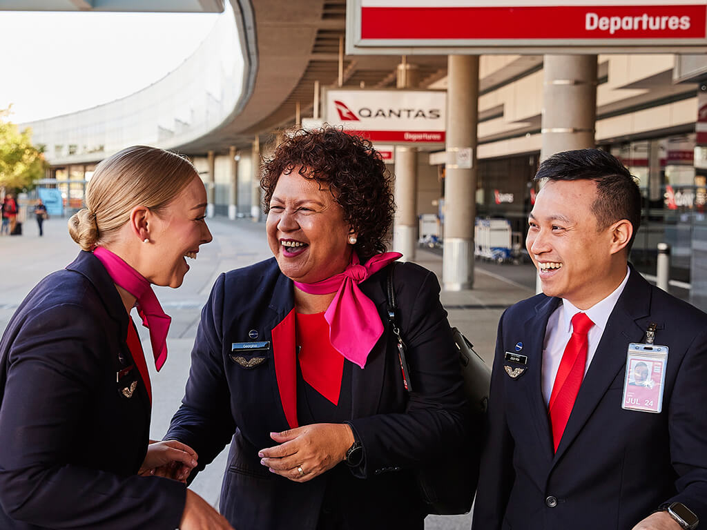 Three Qantas Airport Customer Service employees engaging with passengers outside the terminal, highlighting friendly and professional customer interactions