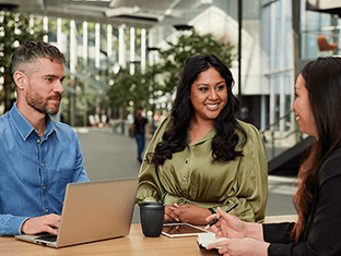 Three Qantas employees conversing with a laptop and coffee in a modern workspace, showcasing a collaborative and inclusive environment