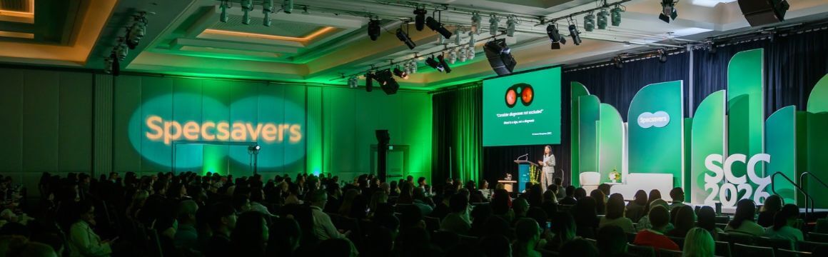 A picture of a conference hall with green lighting, and audience and a person standing on stage