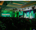 A picture of a conference hall with green lighting, and audience and a person standing on stage