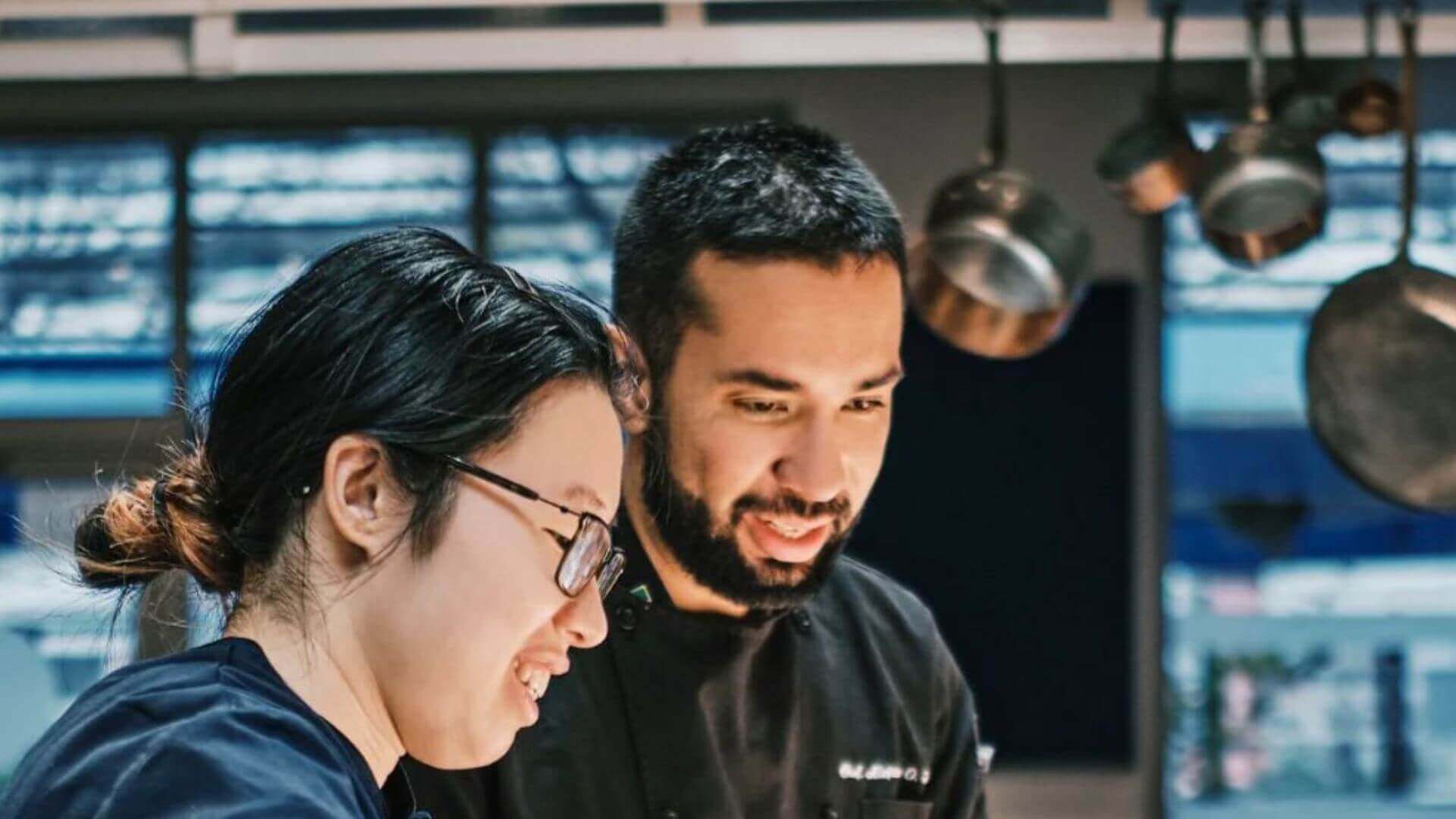 Deux personnes dans une cuisine, avec des casseroles et des poêles en arrière-plan.