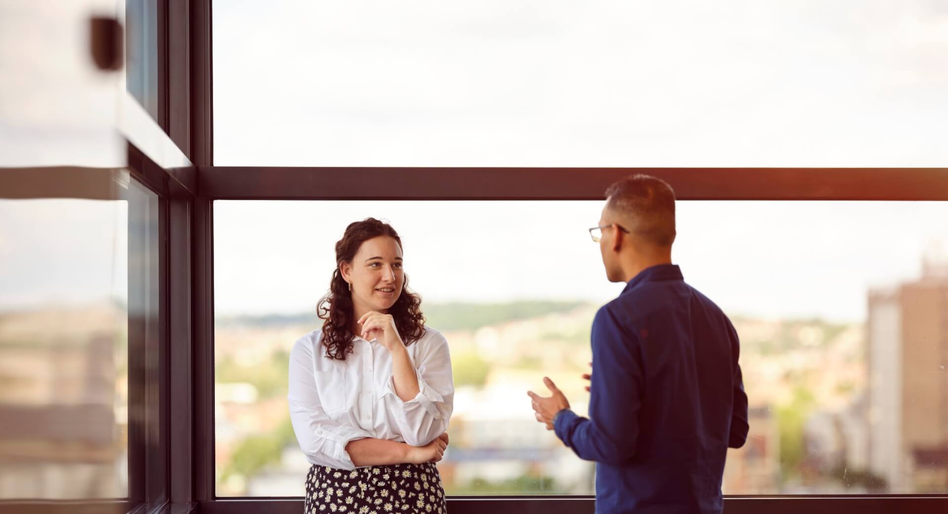 A woman facing the camera talking to a man by a big window in an office