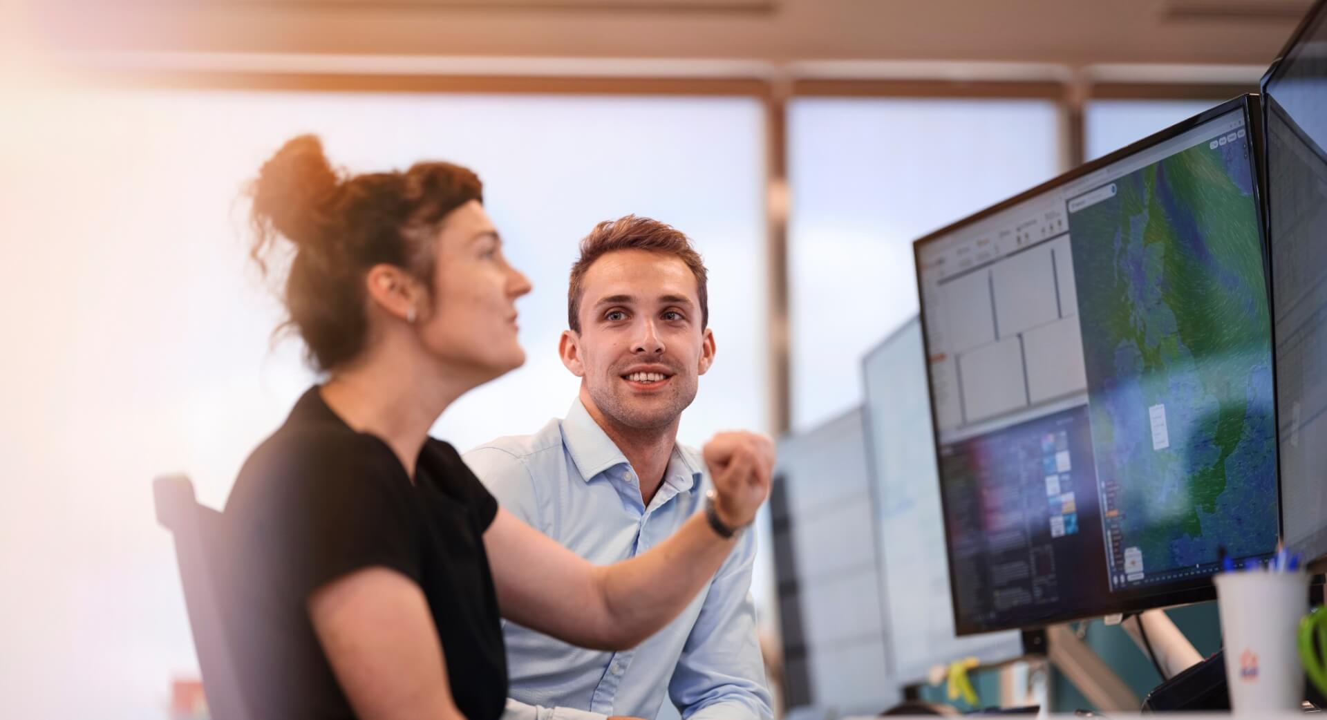 A woman and man chatting in the office. The focal point is the man who is looking at the woman. The woman is looking at a computer screen with her arm raised