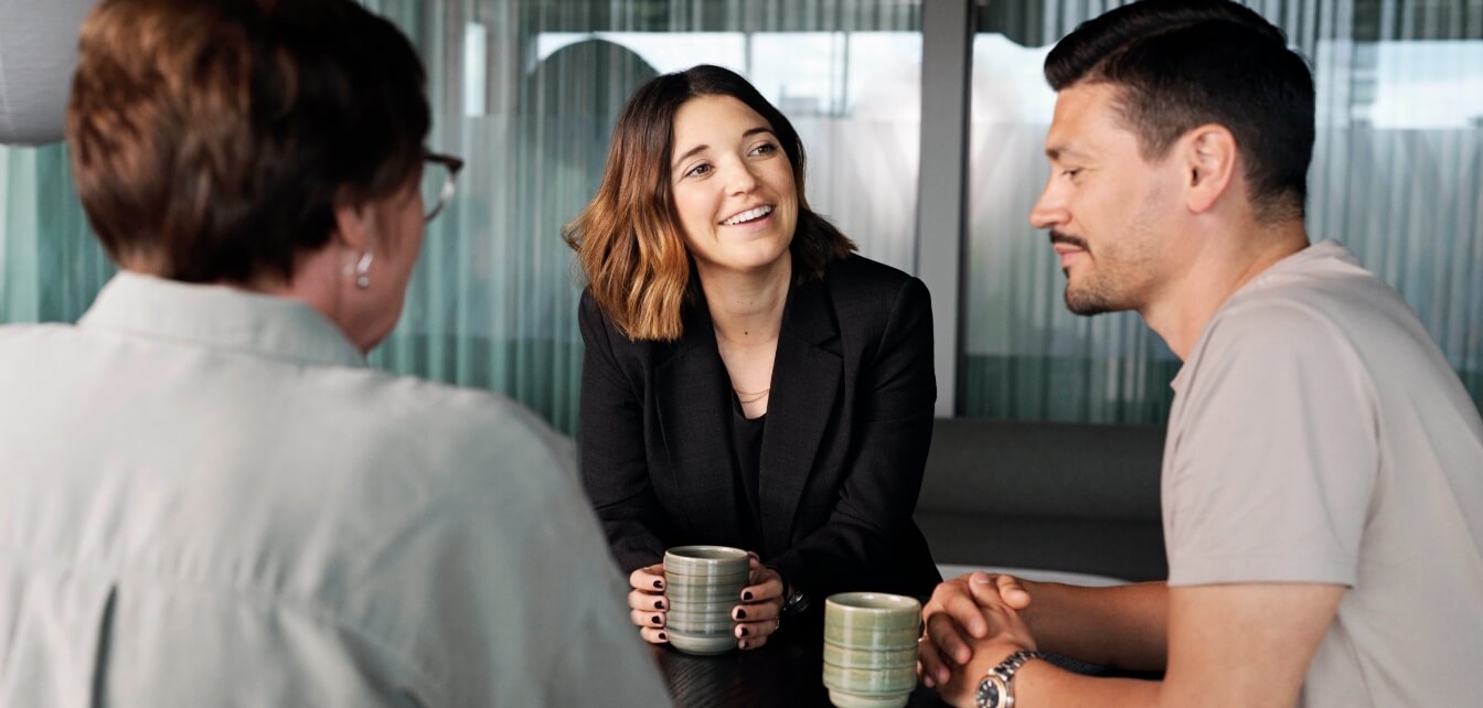 A woman smiling while talking to two people at a table, holding a coffee cup.