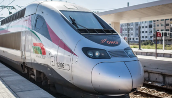 Um elegante e moderno trem de alta velocidade na plataforma de uma estação. O trem tem um design aerodinâmico com exterior prateado e detalhes em vermelho, verde e branco. A frente do trem apresenta o logotipo da ONCF (Office National des Chemins de Fer), a operadora ferroviária marroquina. A plataforma da estação está parcialmente coberta por um dossel e há prédios visíveis ao fundo.