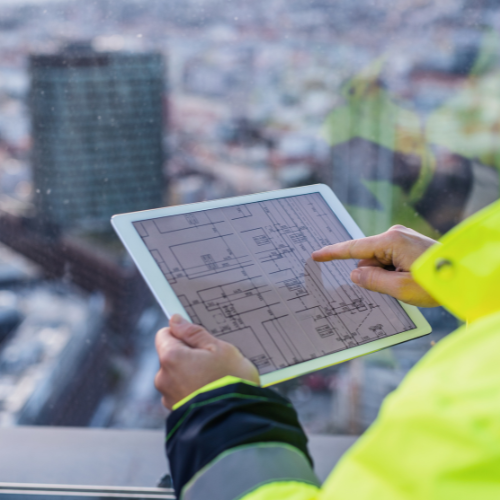 Um trabalhador da construção civil em uma jaqueta de alta visibilidade usando um tablet para revisar planos arquitetônicos, com uma paisagem urbana borrada visível através de uma janela ao fundo.