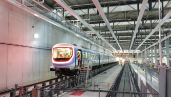 Um trem de metrô moderno dentro de uma instalação de manutenção bem iluminada do Grand Paris Express. O trem está posicionado nos trilhos com uma plataforma de manutenção e uma escada ao lado. A instalação apresenta tetos altos com vigas de metal e vários equipamentos técnicos, indicando uma infraestrutura de última geração para o transporte urbano.