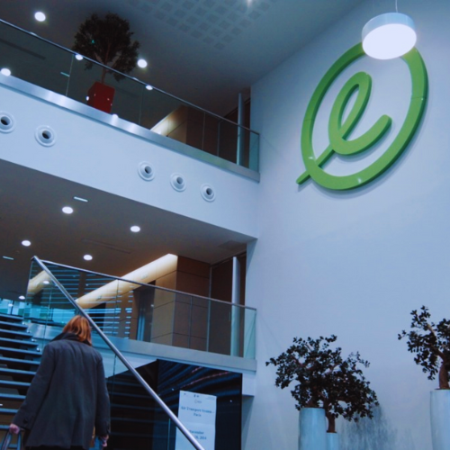 Interior de um escritório moderno com uma escada que leva a um andar superior com grades de vidro. Uma pessoa de cabelos ruivos e casaco cinza está subindo as escadas. A parede do lado direito exibe um grande logotipo verde “e”. A área é bem iluminada com luzes no teto e tem vasos de plantas, criando uma atmosfera acolhedora e profissional.