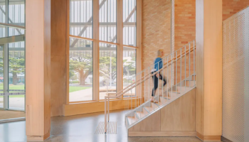 Uma pessoa subindo uma escada dentro de um edifício moderno com grandes janelas do chão ao teto, que é o Cowes Cultural Centre. O interior apresenta detalhes em madeira e um piso de concreto polido. Do lado de fora das janelas, há vegetação e árvores visíveis, criando um contraste entre os ambientes interno e externo.