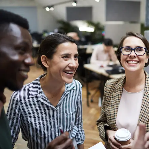 Three people stand together in an office, smiling and chatting. One holds a cup of coffee. Desks and a blurred colleague are visible in the background.