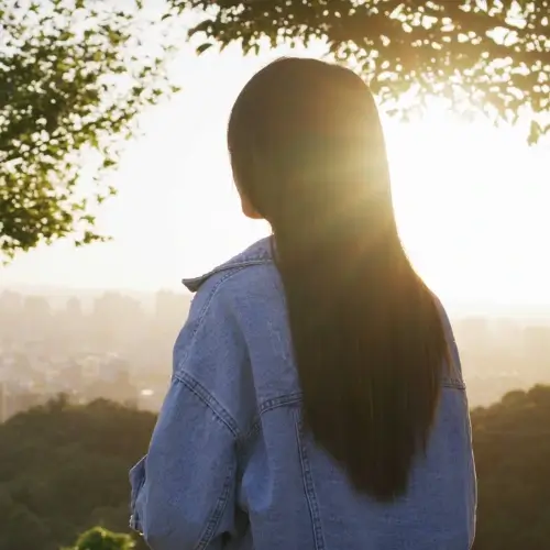 Uma pessoa de cabelos longos vestindo uma jaqueta jeans, em pé ao ar livre e olhando para o horizonte durante o pôr do sol, com uma paisagem urbana visível à distância e árvores emoldurando a cena.