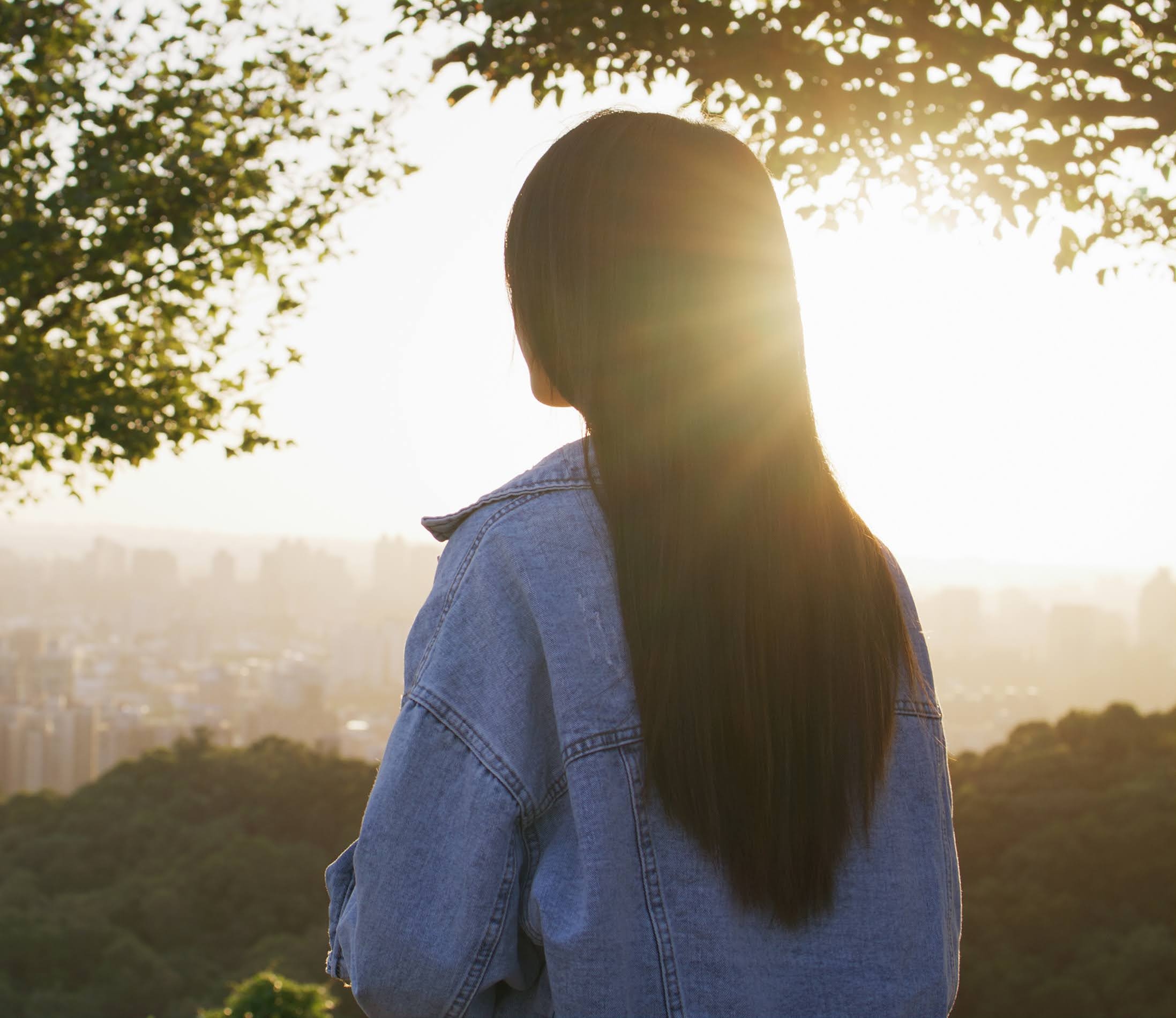 The back of a woman under a tree looking at the sun shining on the city 