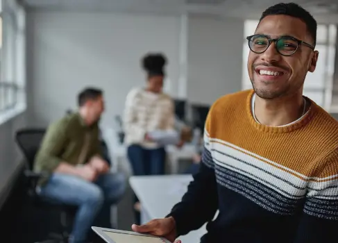  Une personne vêtue d'un pull élégant tenant une tablette dans un environnement de bureau moderne, avec des collègues travaillant en collaboration à l'arrière-plan.