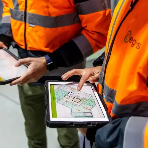 Un ouvrier de la construction portant un gilet de haute visibilité et un casque de protection tient des plans et pointe du doigt un chantier de construction, avec un drone volant au-dessus de lui et du matériel d'arpentage au premier plan.