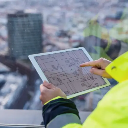 Un ouvrier du bâtiment portant une veste de haute visibilité utilise une tablette pour examiner des plans architecturaux, avec un paysage urbain flou visible à travers une fenêtre à l'arrière-plan.
