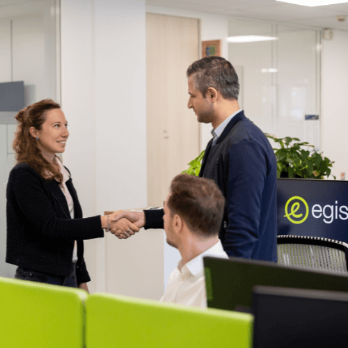 Deux personnes se serrent la main dans un bureau moderne. Le bureau présente des cloisons vertes et un grand panneau avec le logo « egis » bien en évidence. Une troisième personne est assise à un bureau et observe la poignée de main. L'environnement est lumineux et professionnel, avec des plantes en pot qui ajoutent une touche de verdure.