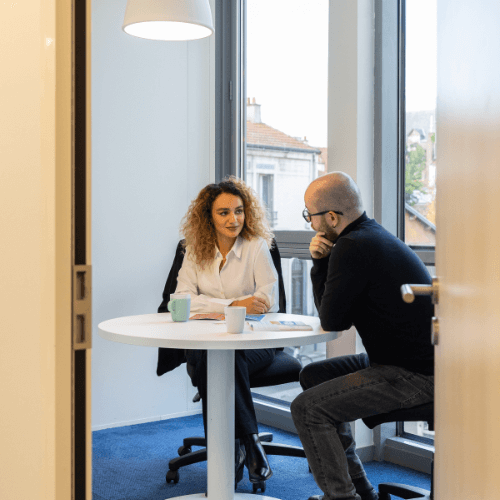 Deux personnes sont assises à une table ronde dans un bureau moderne et discutent. Le bureau est doté de grandes fenêtres qui laissent entrer la lumière naturelle dans la pièce, et une lampe suspendue est accrochée au-dessus de la table. Les deux personnes sont en tenue de travail, et des tasses à café et des documents sont posés sur la table, ce qui indique qu'il s'agit d'une réunion professionnelle. L'atmosphère est lumineuse et propice à des discussions productives.