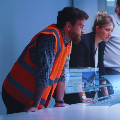 Two individuals, one in a high-visibility vest, are engaged in a discussion while looking at a computer screen displaying architectural plans and technical drawings. They are in a modern office setting with a focus on construction and engineering.