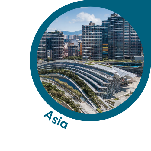 A circular image with a teal border showing a modern cityscape in Asia. The image features tall buildings and skyscrapers, with a distinctive curved architectural structure in the foreground, probably a convention centre or transport hub. The background includes other urban infrastructure and distant mountains under a clear sky. The text ‘Asia’ is written in teal, curving along the bottom left edge of the circle, indicating the Asian context of the scene.