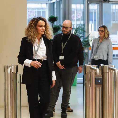 Three people with lanyards walk through a security turnstile in an office building. The front person wears a black suit.