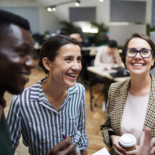 Three people stand together in an office, smiling and chatting. One holds a cup of coffee. Desks and a blurred colleague are visible in the background.