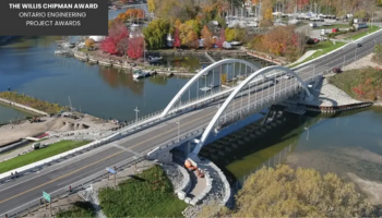  An aerial view of the Road 21 bridge with elegant arches spanning over a river. The bridge is surrounded by lush greenery and autumn-colored trees, with a few boats docked along the riverbank. The image features a caption in the top left corner that reads "The Willis Chipman Award, Ontario Engineering Project Awards," highlighting the bridge's recognition for engineering excellence.