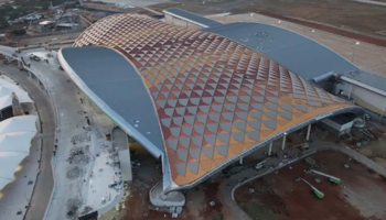 An aerial view of the Trichy Airport, a large, modern building with an undulating, geometric-patterned roof The roof features a mix of red, orange, and grey colors, creating a striking visual effect. The building is surrounded by construction areas, with some vehicles and equipment visible. The landscape around the building includes paved roads and open fields, indicating an ongoing development project.